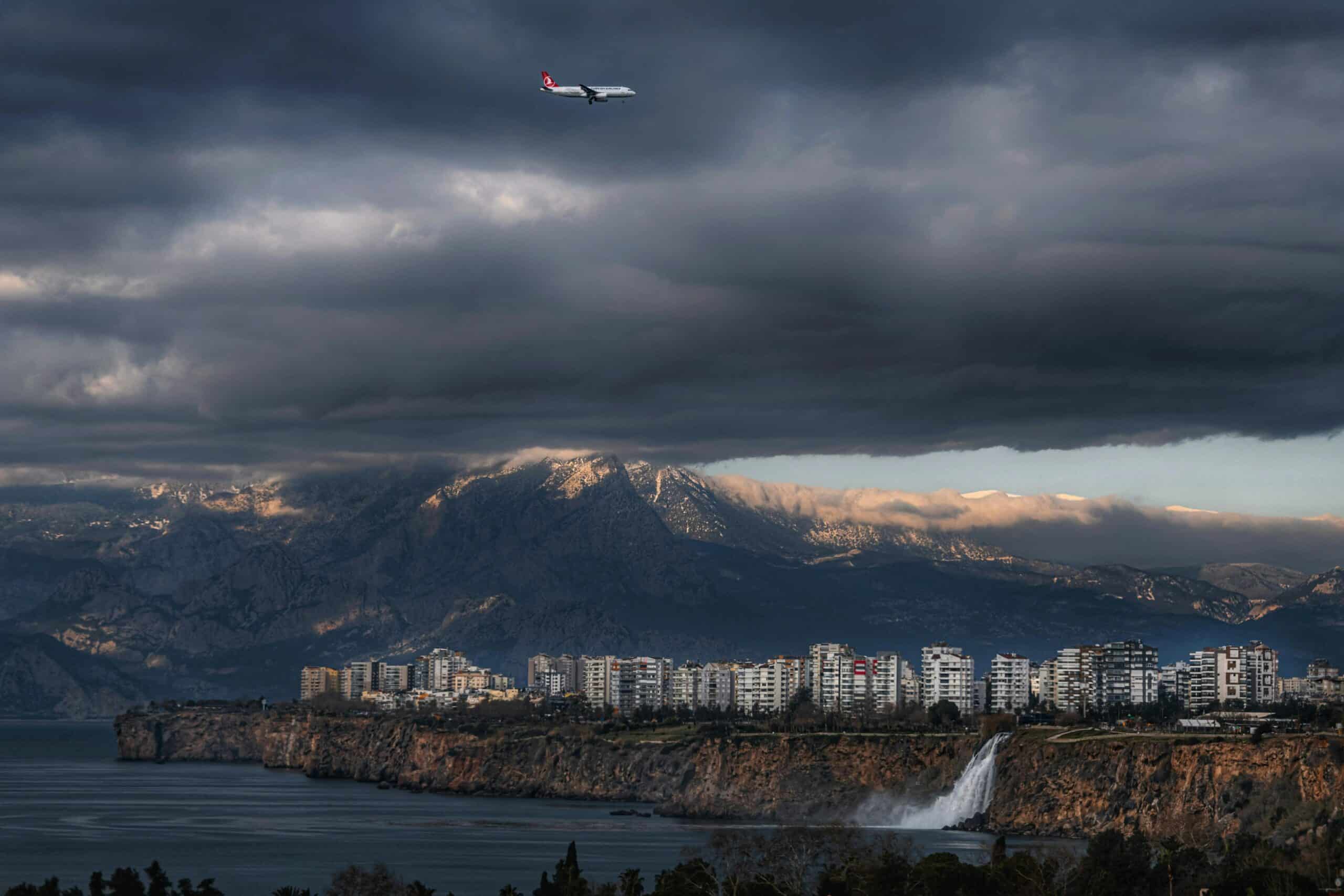 Stunning Antalya cityscape featuring waterfalls, mountains, and an aircraft under a stormy sky.